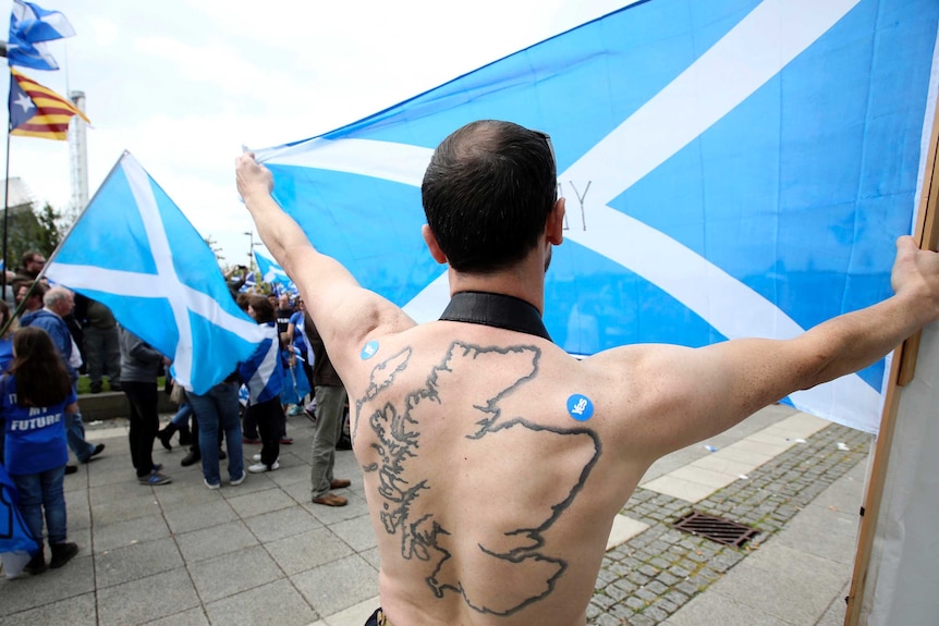 A man with a Scottish map tattooed on his back holds up a Scottish flag.