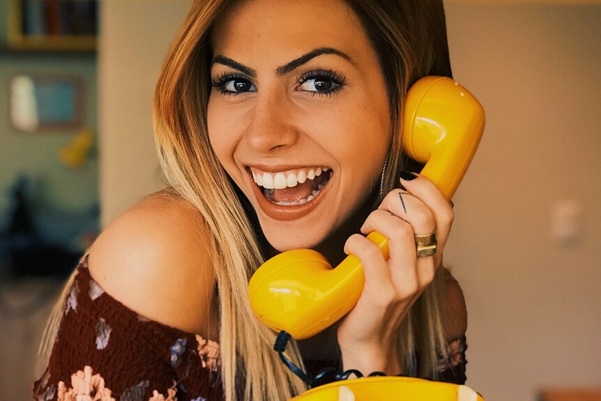 A woman holding a yellow telephone handle to her ear and calling somebody rather than using social media to communicate.