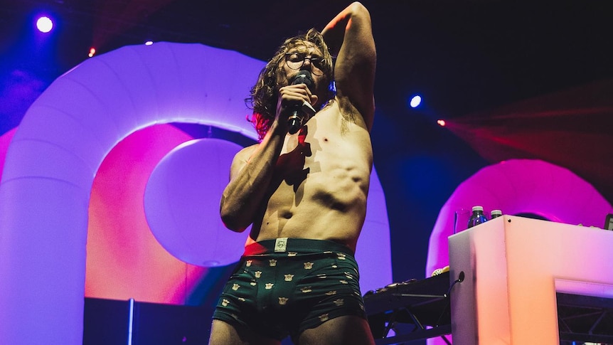 man in boxer shorts with long hair and spectacles arm behind his head on stage singing into microphone