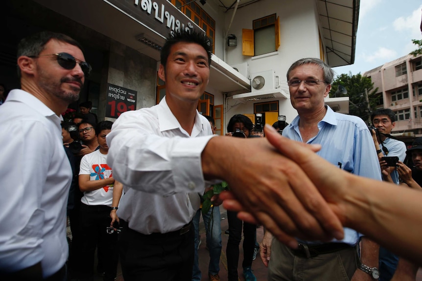 A man in a white shirt shakes hands with someone off camera as two Western diplomats stand side by side.