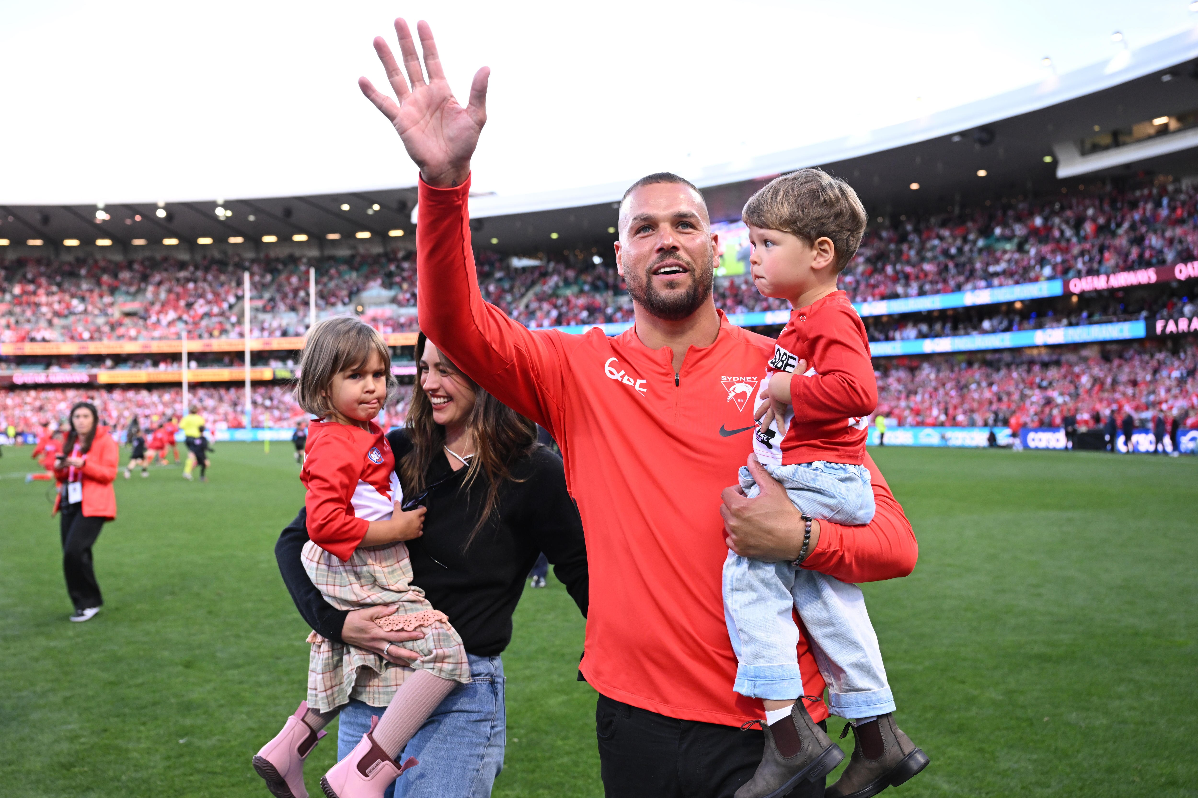 Retired Sydney Swans star Lance Franklin waves to the crowd while holding his son in his arms, his wife and daughter near him.