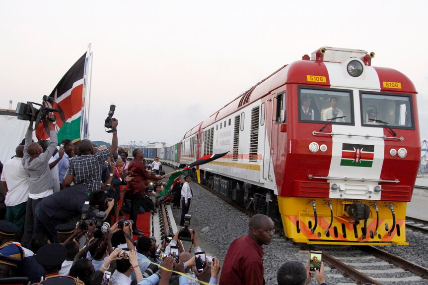 A red, white and yellow cargo train travels along a railway past an enthusiastic crowd waving Kenyan flags.
