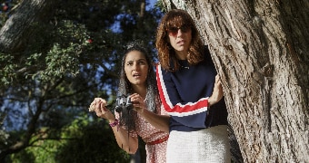 Colour still of Madeleine Sami and Jackie van Beek in 2018 film The Breaker Upperers with binoculars peering from behind a tree.