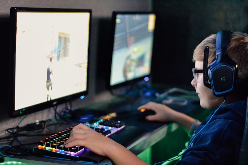 A teenage boy wearing glasses and a gaming headset looks at a computer screen where a game is showing