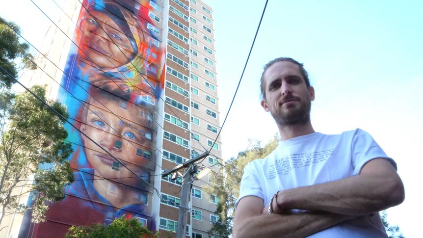 Artist Matt Adnate stands front of a mural of boy and woman wearing a headscarf painted on the side of an apartment block.