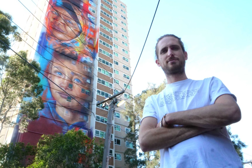 Artist Matt Adnate stands front of a mural of boy and woman wearing a headscarf painted on the side of an apartment block.