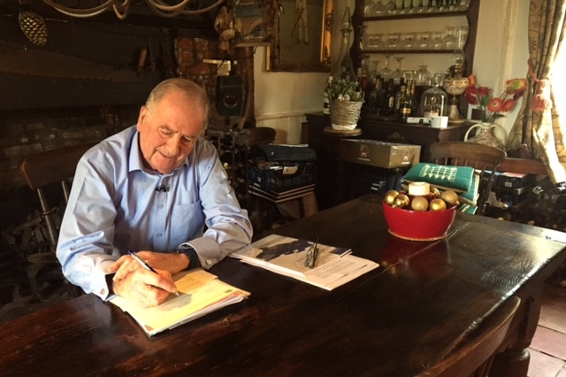 Roger Gale sits writing at a kitchen table. January 2017.