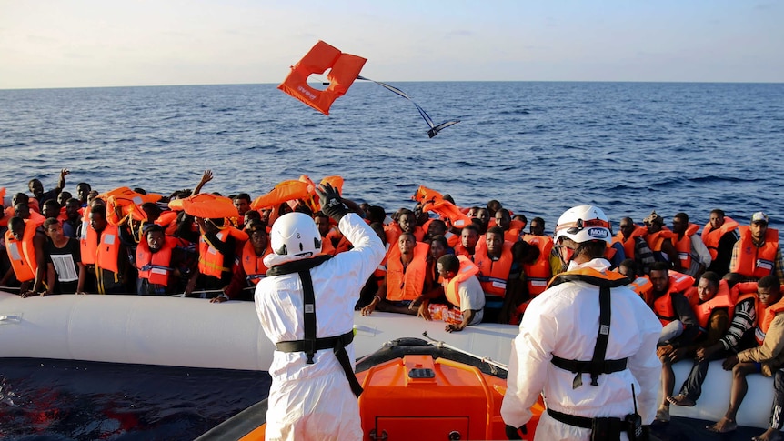 A life jacket is thrown by rescuers to asylum seekers on a rubber dinghy. An expanse of water from the ocean behind them.