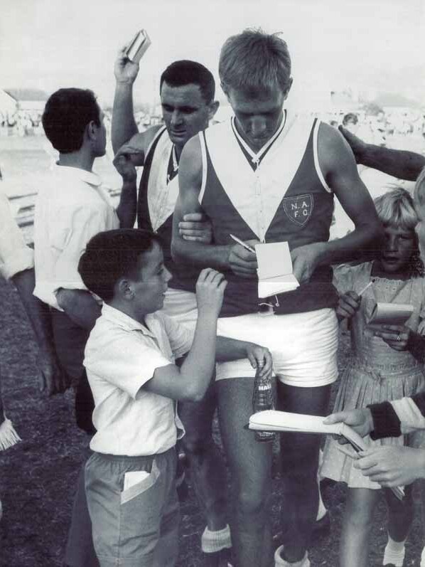 A North Adelaide SANFL footballer stands with a pen in his hand signing a notebook for young boy after a game.