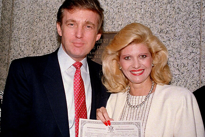 A red-haired man and a blonde woman stand together. He's in a dark suit, she's wearing a cream jacket and holds a certificate