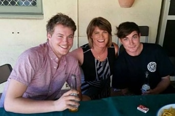 A mother sits at an outdoor table with her two sons on either side, all posing for a photo.