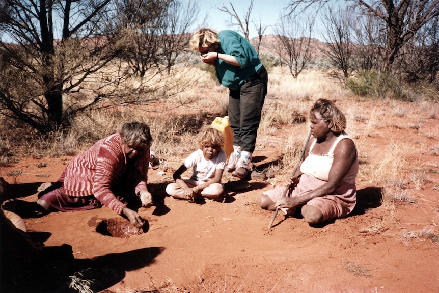 Two women and a child sitting on the ground while one of the woman makes a clay slurry.