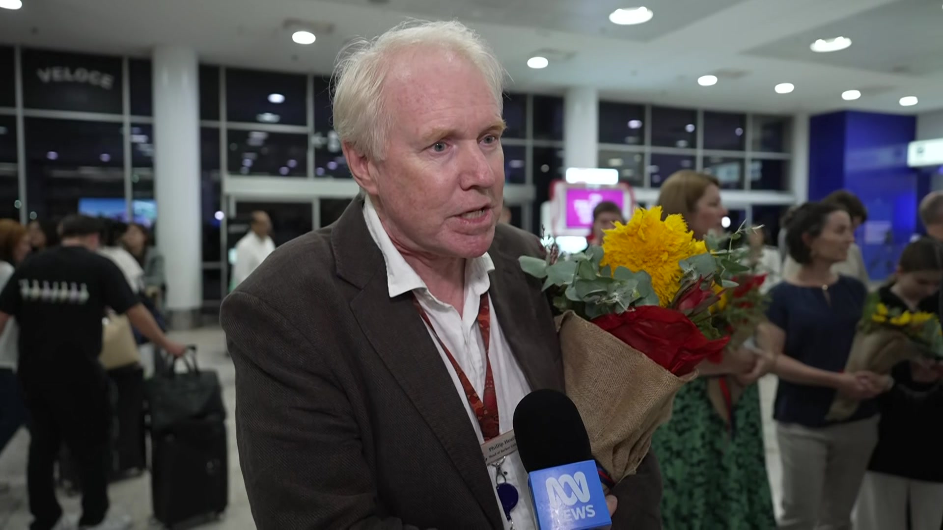 Un hombre de mediana edad con un traje marrón sostiene flores mientras habla con un periodista en un aeropuerto.