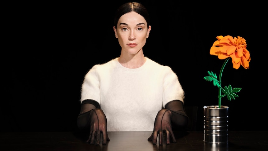 St Vincent sits in darkness, wearing white jumper and black gloves, at a table with an orange flower in a tin can