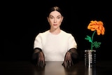 St Vincent sits in darkness, wearing white jumper and black gloves, at a table with an orange flower in a tin can