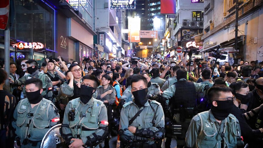 People are hemmed in on a narrow Hong Kong street by a row of police as neon lights on buildings shine brightly.