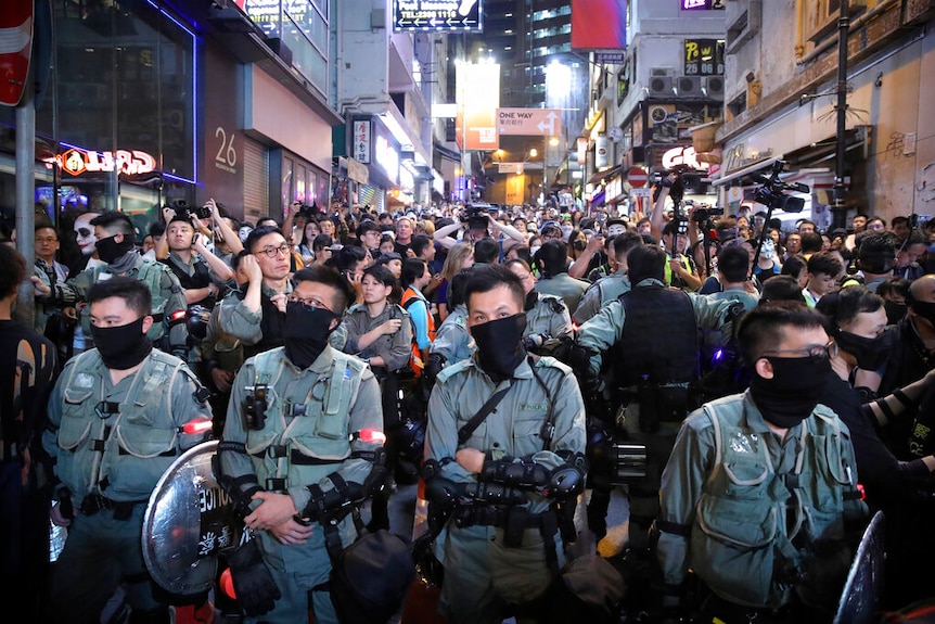 People are hemmed in on a narrow Hong Kong street by a row of police as neon lights on buildings shine brightly.