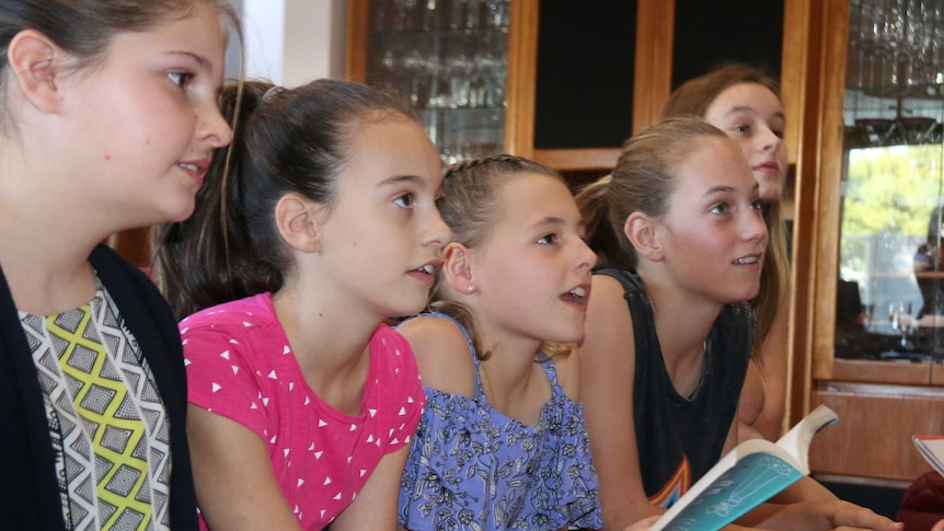 Five girls sit on a couch looking at a friend discussing a book