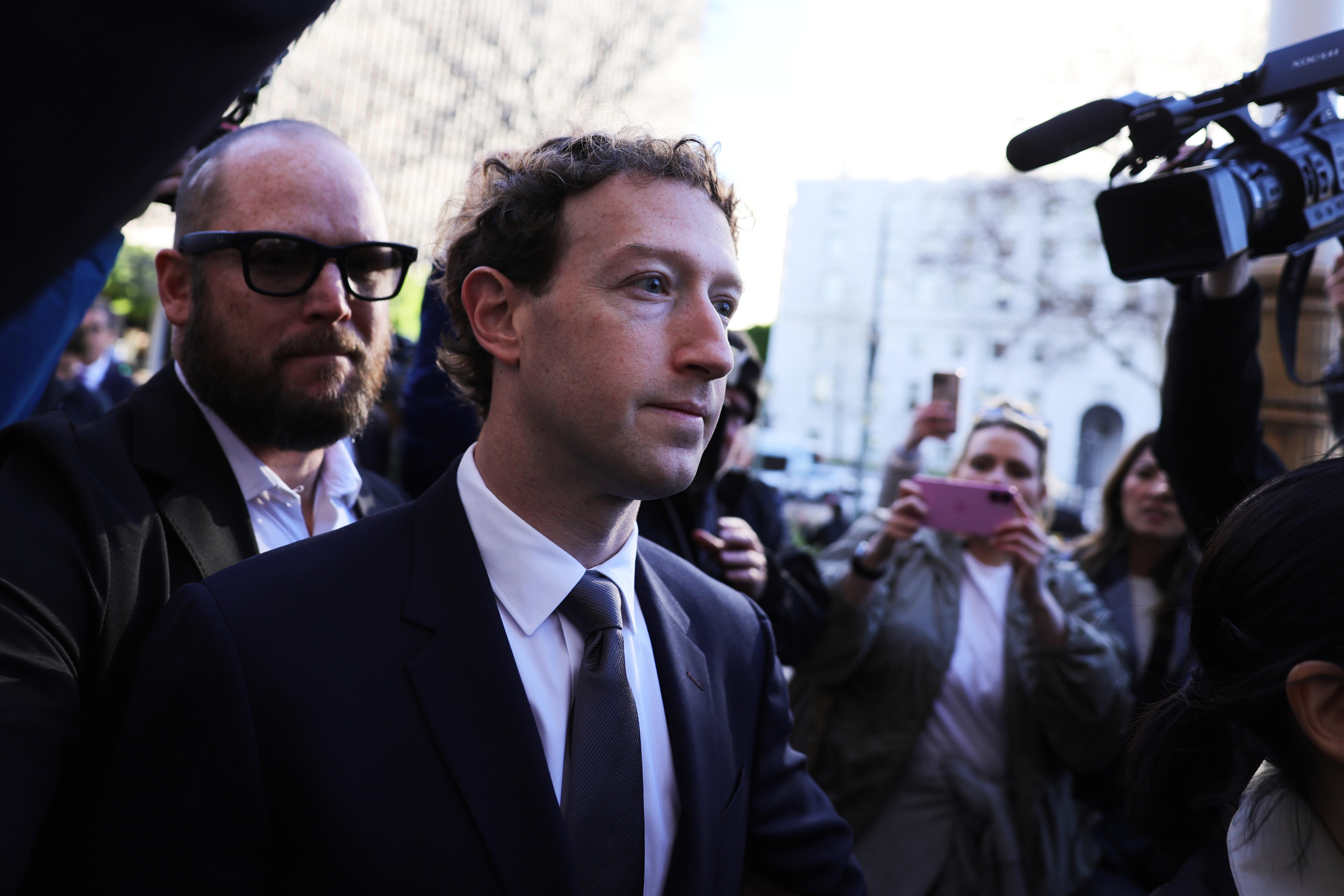 Mark Zuckerberg wearing a suit with several people pointing their phones and a camera at him.