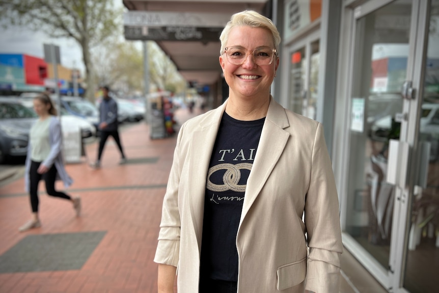 A woman with blonde hair and a cream blazer smiles for a portrait photo while standing on a street.