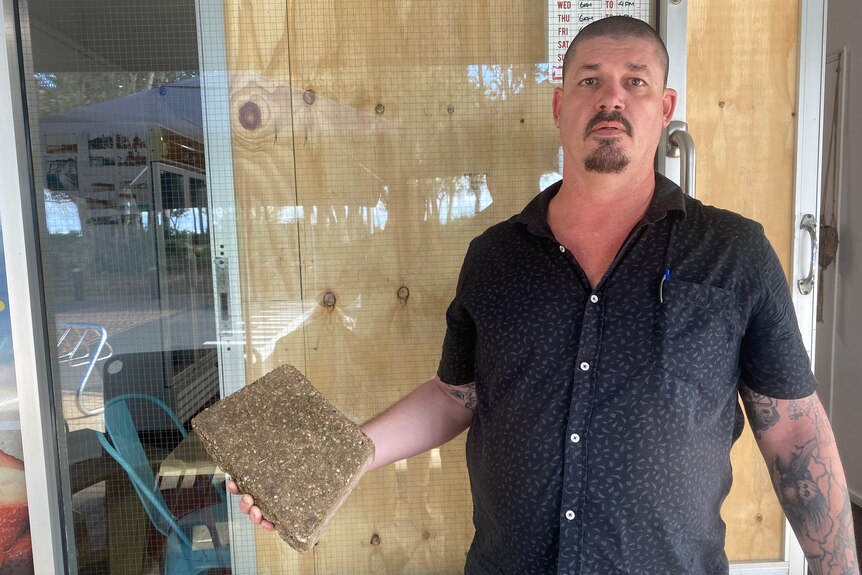 Middle aged man stands in front of shop front with brick in hand.