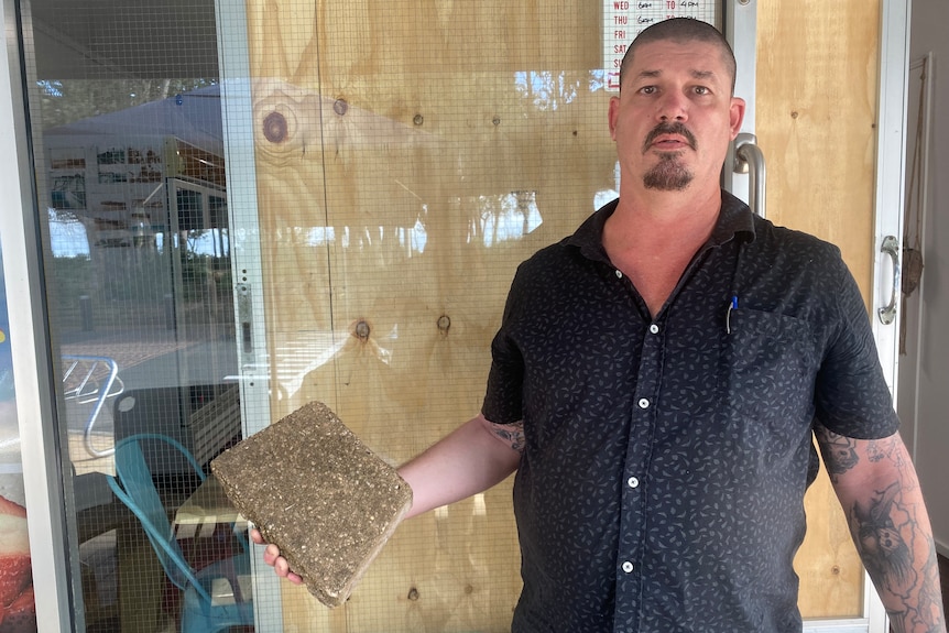 Middle aged man stands in front of shop front with brick in hand