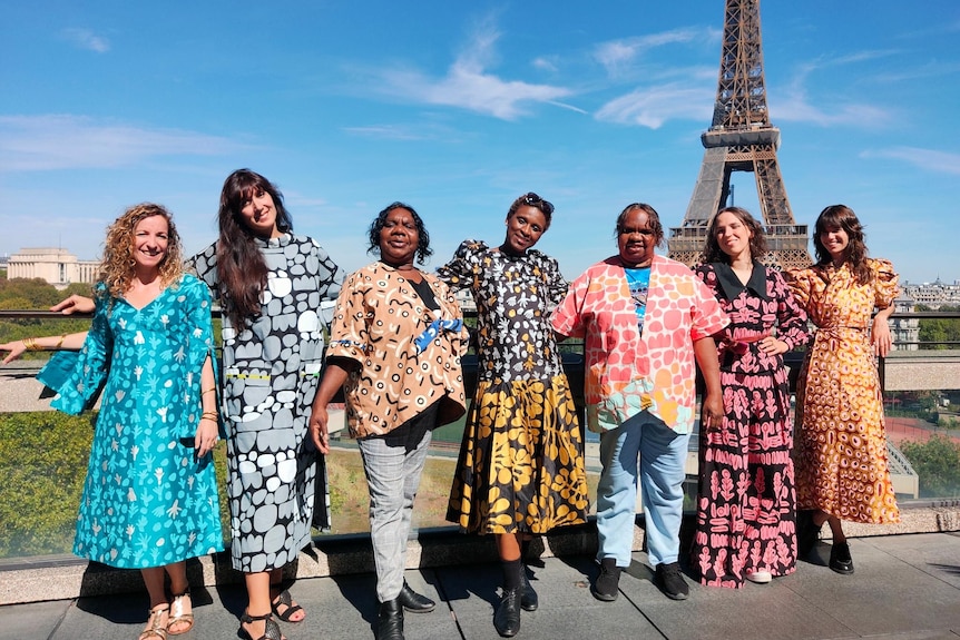 Seven women wearing colourful Indigenous textiles smile and link arms in front of the Eiffel Tower