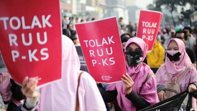 Three women wearing hijabs with plastered mouths holding papers that read 'reject the TPKS Bill' in a protest.