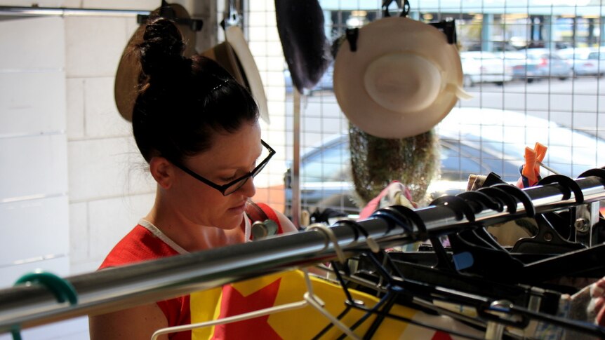 A woman wearing glasses flicks through racks of clothes in an op shop.