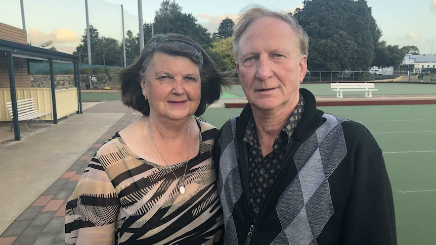 A man and a woman stand together looking directly at the camera. Bowling greens are in the background.