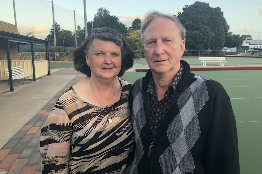 A man and a woman stand together looking directly at the camera. Bowling greens are in the background.