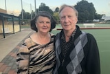 A man and a woman stand together looking directly at the camera. Bowling greens are in the background.