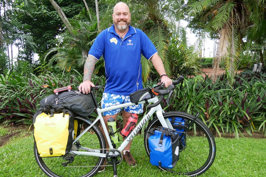 A man in a blue shirt stands behind his bicycle, in front of trees in a park, smiling.