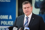 A middle aged man speaks directly to camera in front of press microphones in a neat suit with the background out of focus.