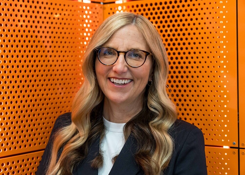 Middle-aged woman who has long blonde hair and is wearing glasses in front of an orange backdrop