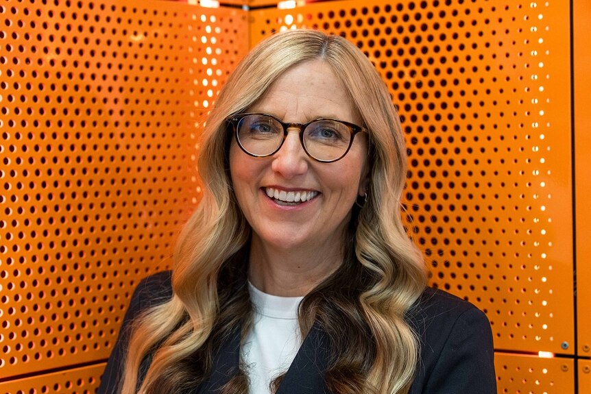 Middle-aged woman who has long blonde hair and is wearing glasses in front of an orange backdrop