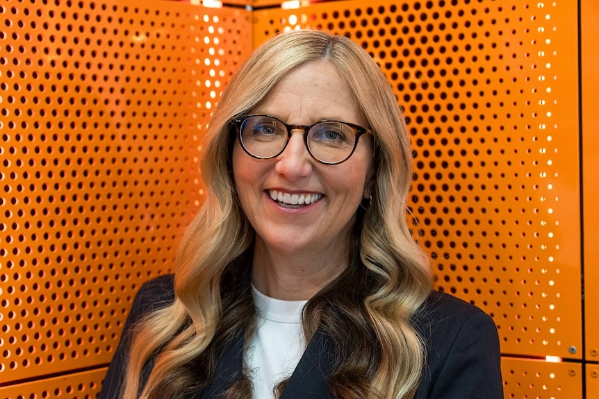 Middle-aged woman who has long blonde hair and is wearing glasses in front of an orange backdrop
