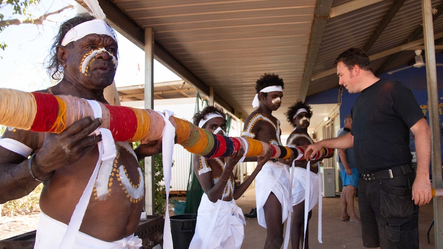 A man in dark shirt and shorts stands with four brightly-painted Indigenous Australians holding a pole