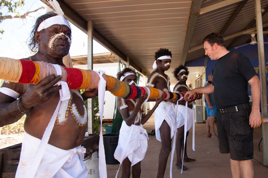 A man in dark shirt and shorts stands with four brightly-painted Indigenous Australians holding a pole