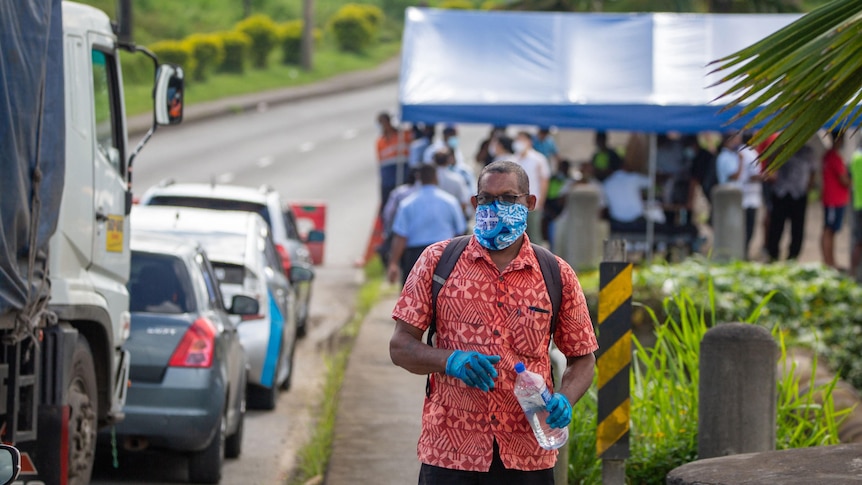 A man in a mask and gloves stands alongside a line of cars with a checkpoint in the background.