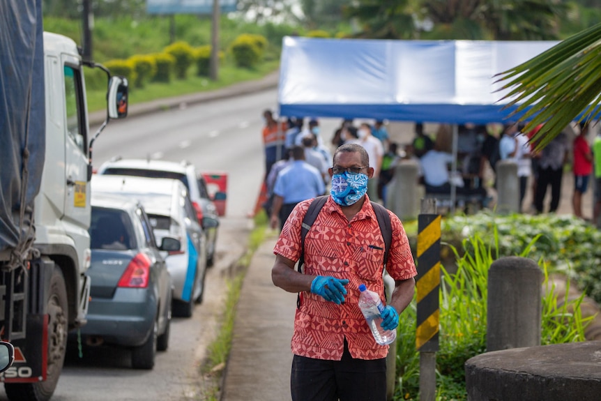 A man in a mask and gloves stands alongside a line of cars with a checkpoint in the background.