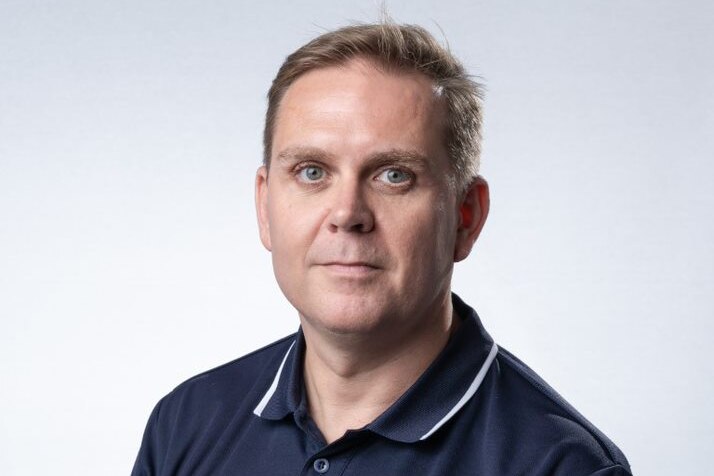 A man wearing a navy shirt, light brown hair and eyes.