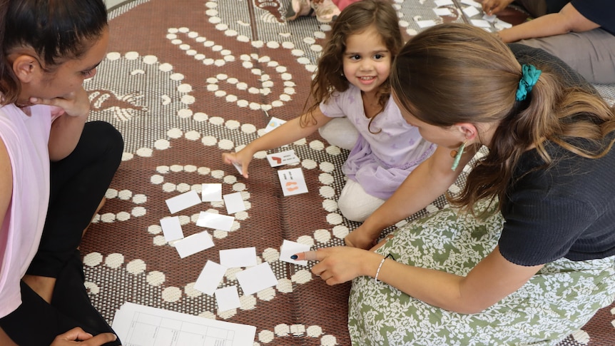 A little girl sits on a rug on the floor with two women. They are looking at flash cards