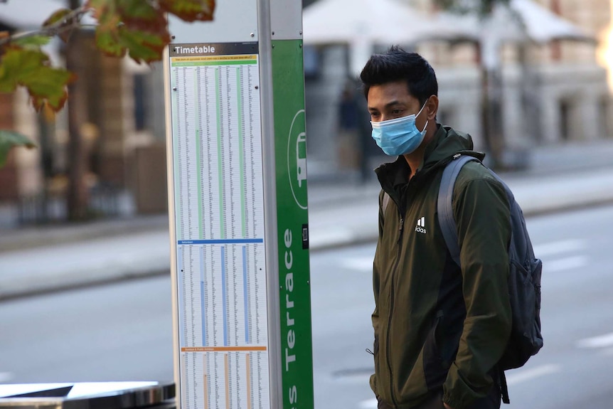 A man wearing a face mask and carrying a backpack stands in the Perth CBD looking at a bus timetable.