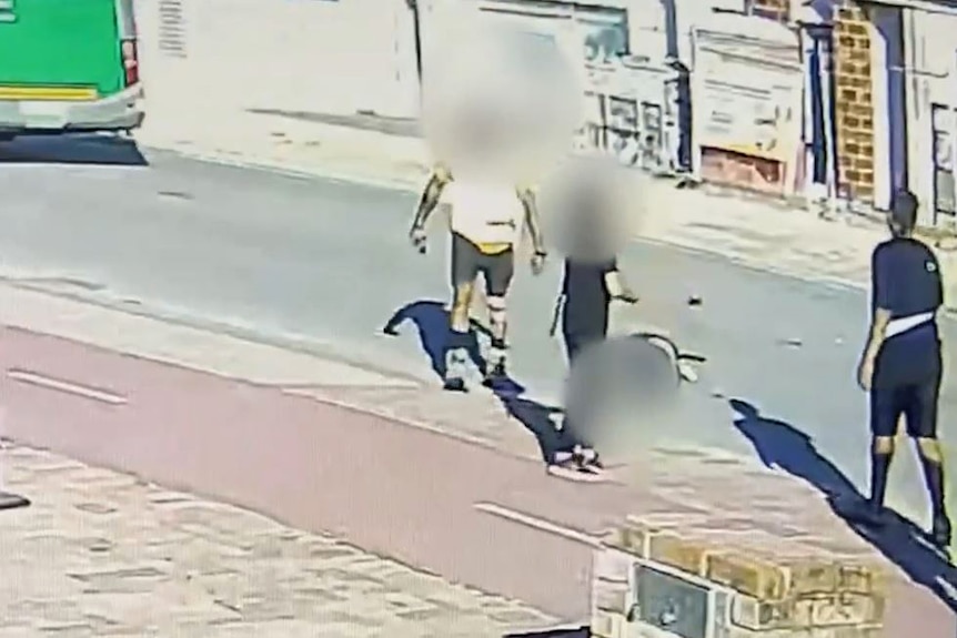 A still image taken from CCTV showing three teenagers standing on a street near a man on the ground, with identities blurred.