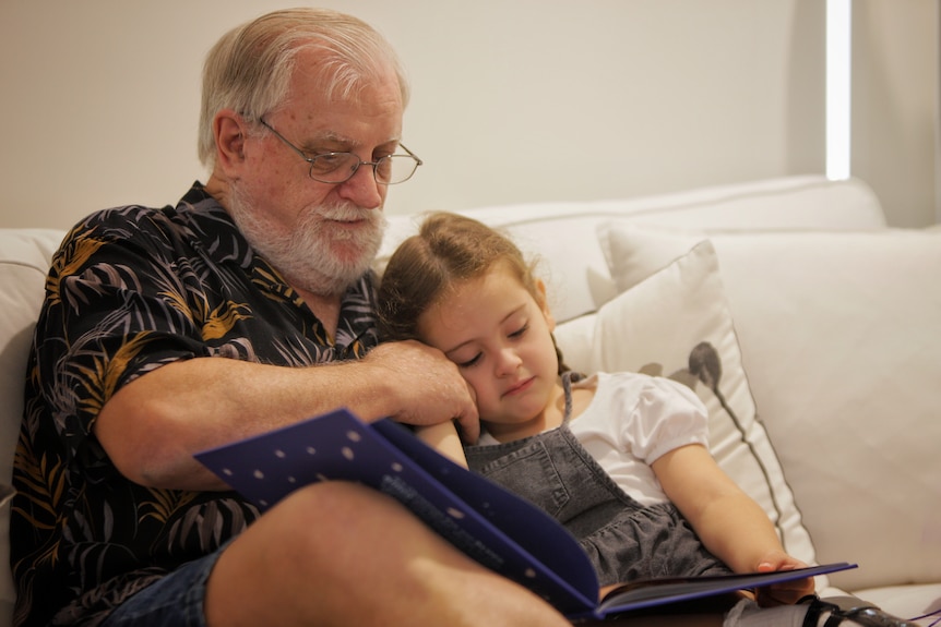 Older man sitting on a couch going through a book with his granddaughter.