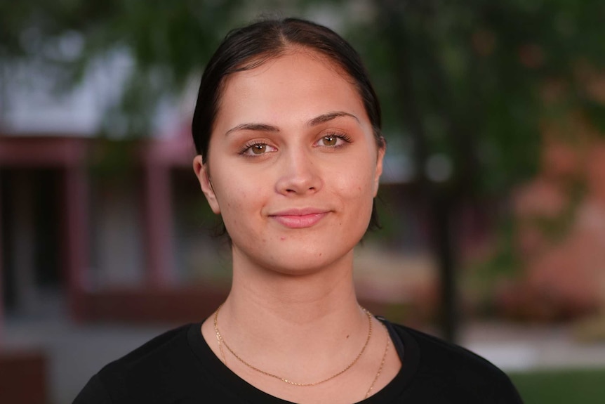 Amishah wears a black t-shirt. She smiles at the camera for the mid-shot. Her hair is pulled back.