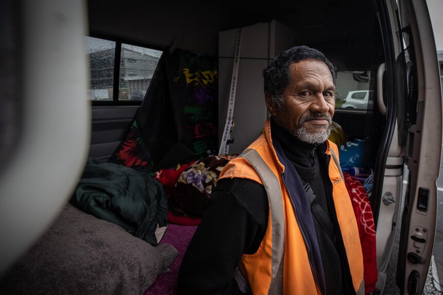 Joseph wears a high vis jacket and sits on the edge of his van. A fridge and blankets can be seen in the back