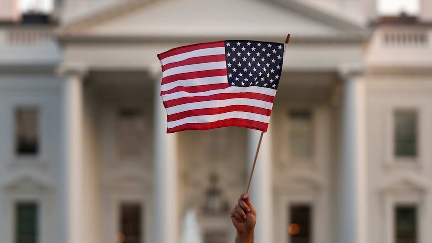 a united states flag is held up by a hand with the White House out of focus behind it
