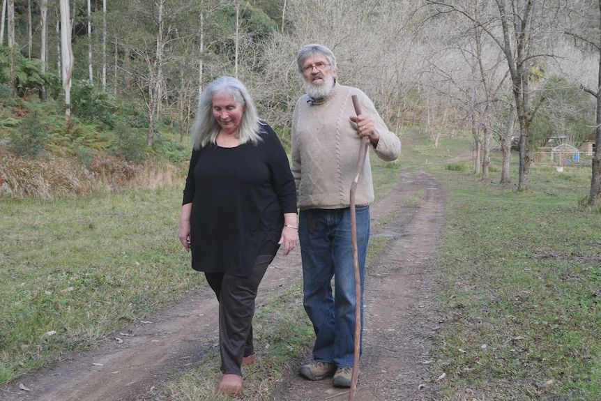 A couple in their 60s walking along a bush driveway the man has a stick to assist walking.